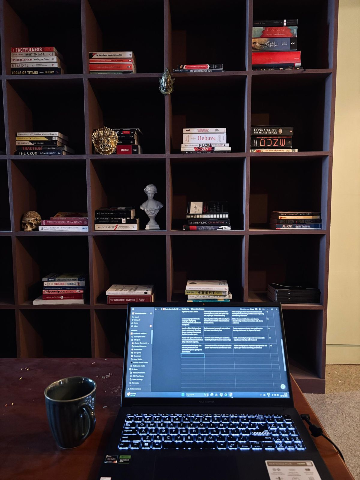 A laptop and a coffee mug on a desk in front of shelves of books